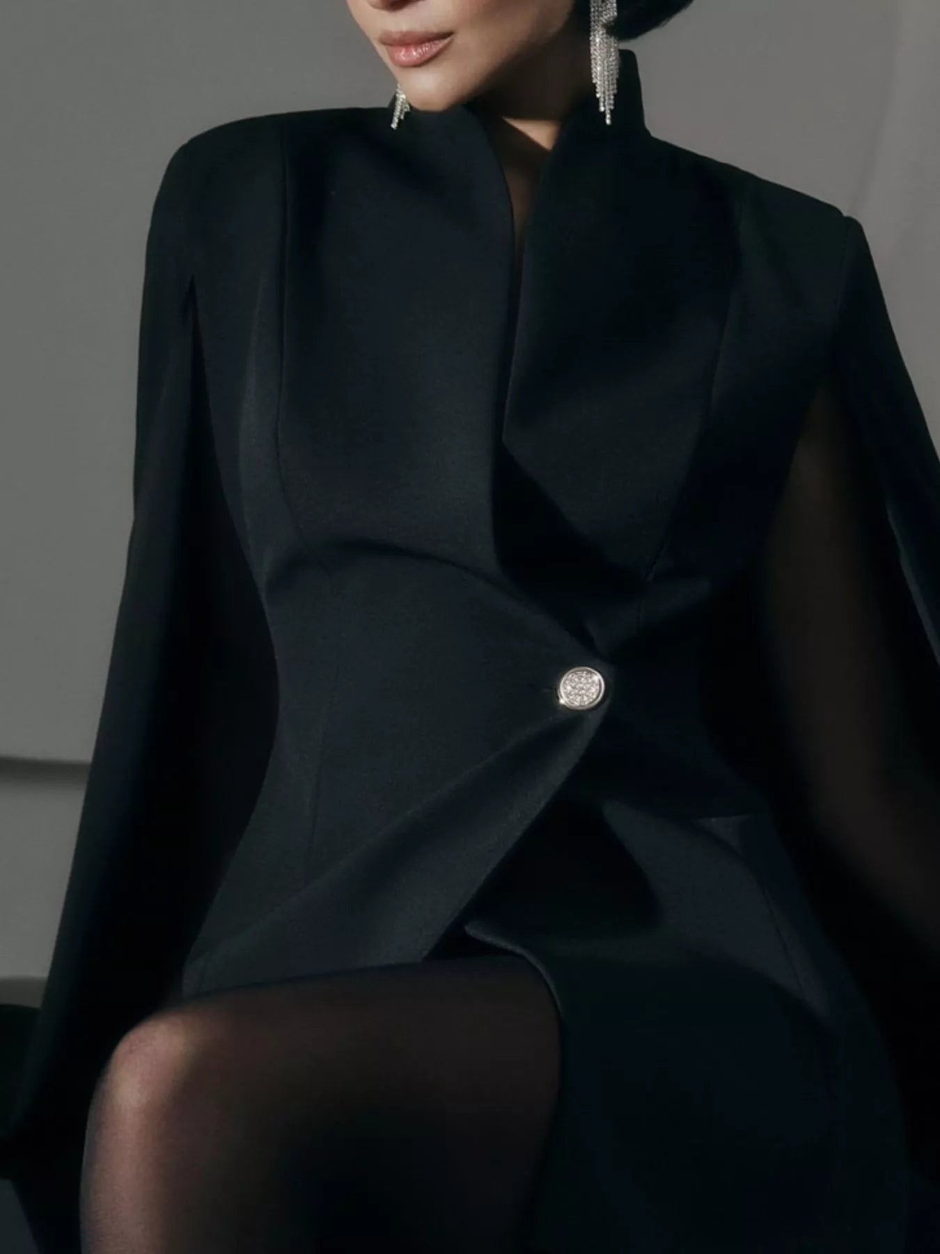 Black fitted mini blazer dress with cape sleeves and one-button front; seated pose